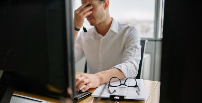 An exhausted professional rubbing his forehead at his desk in front of two monitors