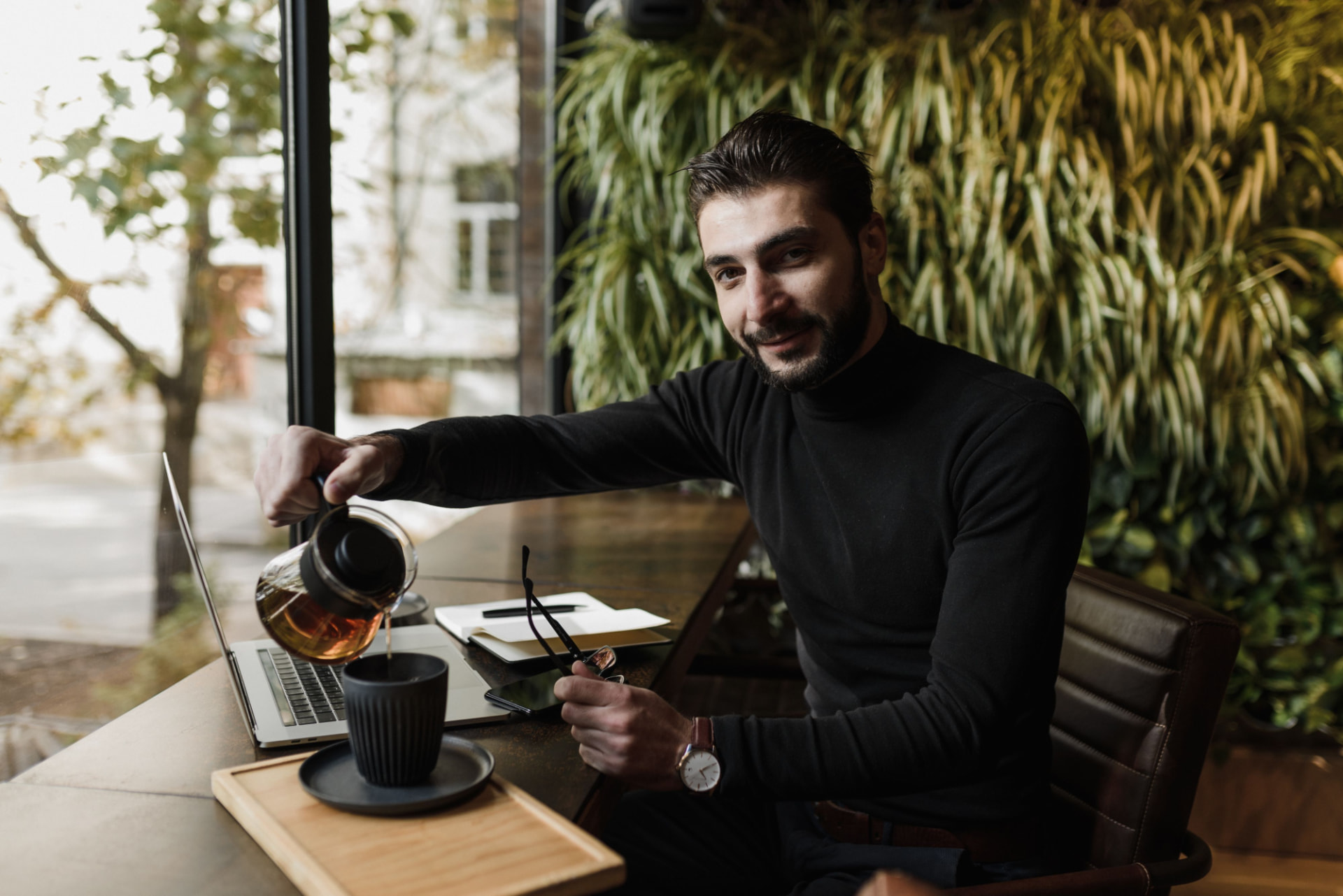 A professional pouring herbal tea beside his laptop in a plant-filled café